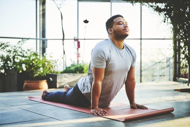 An adult man holds the full cobra pose in a sunroom with plants behind him.